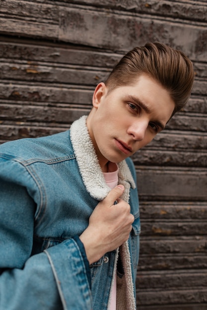 Porträt junger Mann mit ernstem Gesicht mit trendiger Frisur in modischer blauer Jeans-Freizeitjacke in der Nähe der Vintage-Holzwand auf der Straße. Attraktives Kerlmodell in der Modejugendjeanskleidung genießt Spaziergang.