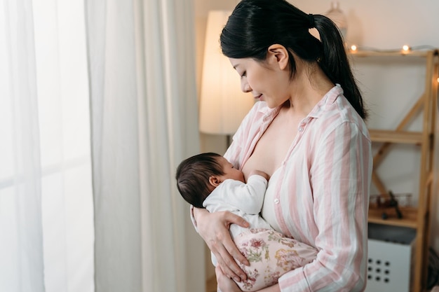 Porträt Koreanische Mutter, die zum ersten Mal in der Nähe des Fensters steht, blickt liebevoll auf die kleine Tochter in ihrer Wiege, während sie sie in einem hellen Wohninterieur stillt.