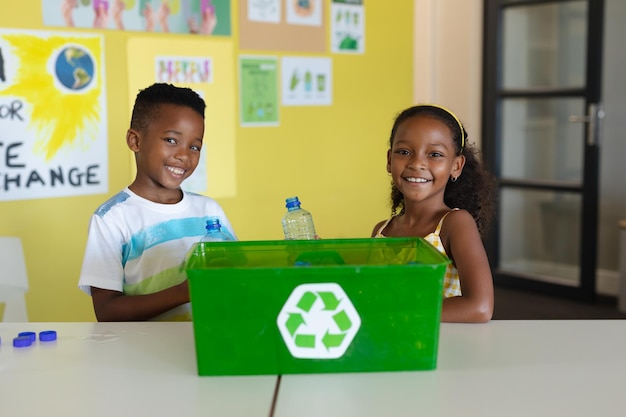 Porträt lächelnder afroamerikanischer Grundschüler mit einem Recyclingbehälter am Schreibtisch in der Schule