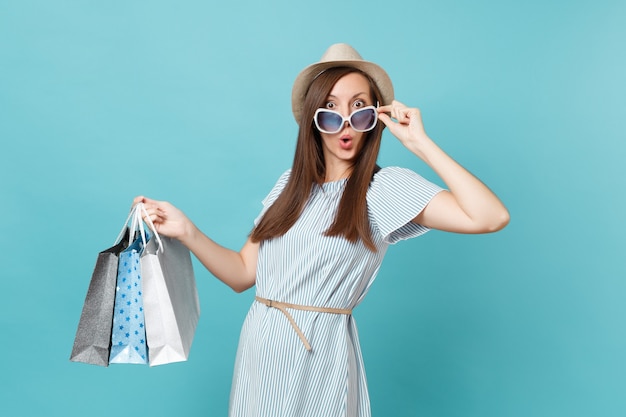 Porträt modische attraktive glückliche Frau im Sommerkleid, Strohhut, Sonnenbrille, die Pakettaschen mit Einkäufen nach dem Einkaufen einzeln auf blauem Pastellhintergrund hält. Kopieren Sie Platz für Werbung.