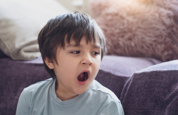 Porträt müde Kind gähnt sitzen neben dem Sofa, schläfriger Junge gähnt und schaut nach unten, Kind hat Allergie während des Wetterwechsels, Kindheit hat Reflexion oder Heuschnupfen von Hausstaubmilbe, Allergien im Kind