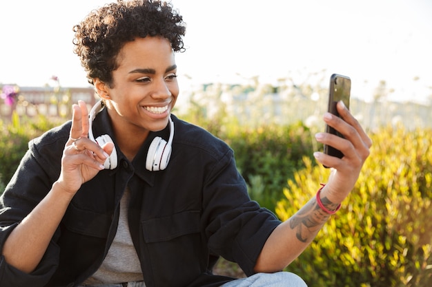 Porträt-Nahaufnahme einer glücklichen Frau mit Kopfhörern, die ein Selfie-Porträt auf dem Handy macht und Frieden zeigt, während sie auf dem Boden sitzt