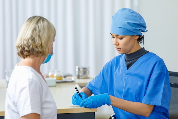 Porträt, Nahaufnahme einer Ärztin in blauer Krankenhausuniform, die auf dem Schreibtisch sitzt und wichtige persönliche Informationen von der kaukasischen älteren Patientin erhält, die eine Gesichtsmaske im Krankenhaus trägt.