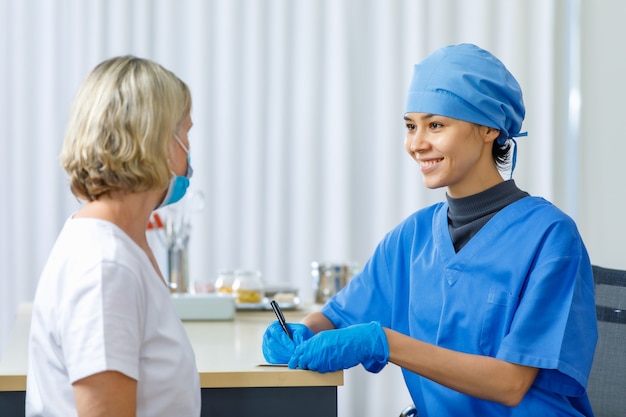 Porträt, Nahaufnahme einer Ärztin in blauer Krankenhausuniform, die auf dem Schreibtisch sitzt und wichtige persönliche Informationen von der kaukasischen älteren Patientin erhält, die eine Gesichtsmaske im Krankenhaus trägt.