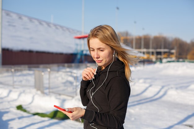 Porträt netten Blondine, die Musik beim ein rotes Telefon in der Hand halten hinunter die Straße gehen hören. Schnee liegt herum.