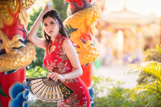 Porträt schön lächelt asiatische junge Frau, die rotes traditionelles chinesisches cheongsam trägt und ein chinesisches Fächern für chinesisches Neujahrsfest am chinesischen Schrein hält