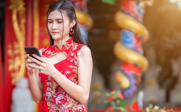 Porträt schön lächelt asiatische junge Frau, die rotes traditionelles chinesisches cheongsam trägt und Nachricht auf Smartphone schreibt