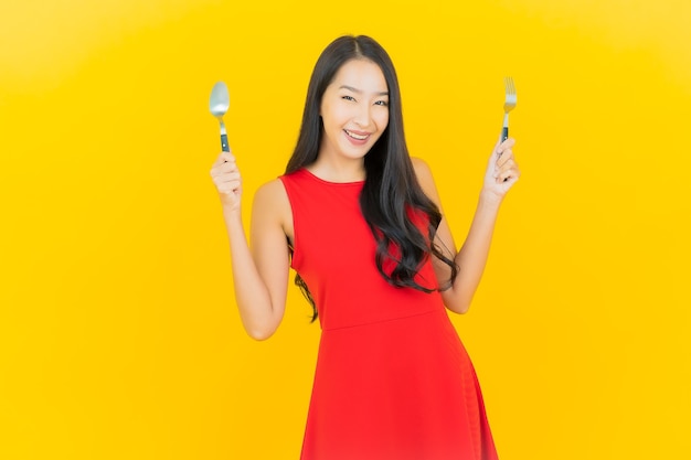 Porträt schöne junge asiatische Frau lächeln mit Löffel und Gabel auf gelber Wand