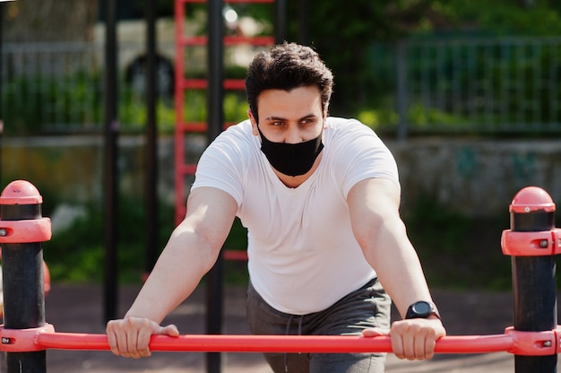 Porträt sportlicher arabischer Mann in der schwarzen medizinischen Gesichtsmaske, die Trainingsübungen im Freiengymnastikplatz während der Coronavirus-Quarantäne tut.