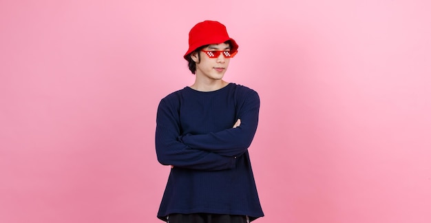 Porträt-Studioaufnahme Asiatisches junges urbanes jugendlich Mode-männliches Model im Street-Style-Outfit Langarm-Shirt roter Eimerhut lustige Brille mit verschränkten Armen lächelnder Blick in die Kamera auf rosa Hintergrund