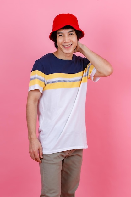 Porträt-Studioaufnahme eines asiatischen jungen gutaussehenden urbanen Teenager-Mode-Männchenmodells im Street-Style-Outfit-T-Shirt mit beigefarbenen Hosen und rotem Eimerhut, der stehend posiert und Blick in die Kamera auf rosa Hintergrund stellt