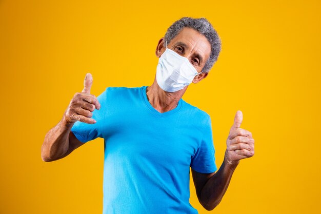 Porträt von älterem Afro mit medizinischer chirurgischer Maske, die in blauem Hemd mit Daumen nach oben steht und Daumen hoch macht.