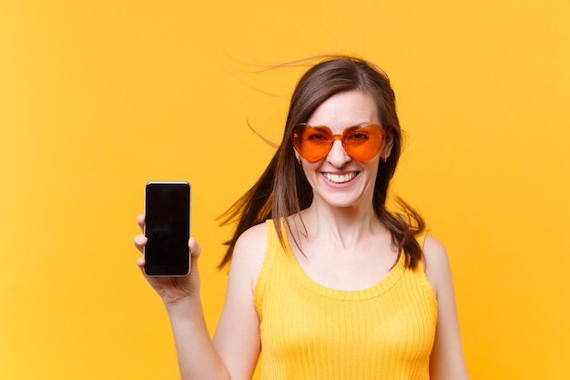 Porträt von aufgeregtem fröhlichem Lachen lustige komische Frau in orangefarbener Brille mit flatterndem Haar halten Handy mit leerem leerem Bildschirmkopierraum einzeln auf gelbem Hintergrund. Werbefläche.