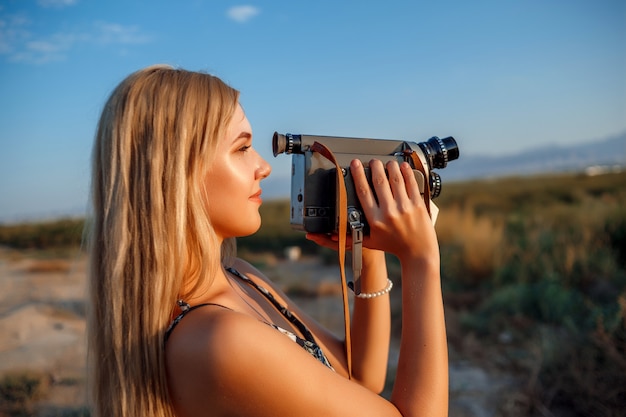 Porträt von Blondinen im Blumendruckkleid mit Weinlesevideokamera auf dem Traubengebiet während des Sonnenuntergangs