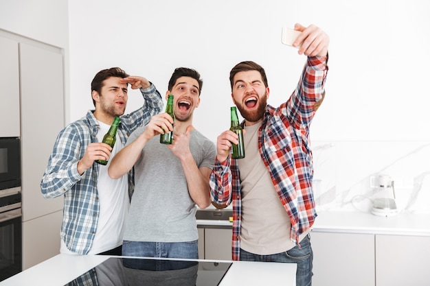 Porträt von drei aufgeregten männlichen Freunden, die feiern, während sie mit Bierflaschen stehen und Selfie drinnen nehmen