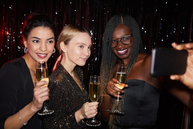 Porträt von drei eleganten jungen Frauen, die Champagnergläser halten und lächeln, während Selfie Foto auf Party machen, mit Blitz geschossen