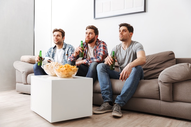 Porträt von drei konzentrierten jungen Männern, die Fußball schauen, während sie zu Hause sitzen, Bier trinken und Snacks essen