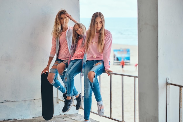 Porträt von drei Schwestern in trendiger Kleidung, die auf einer Leitplanke an einer Meeresküste sitzen.