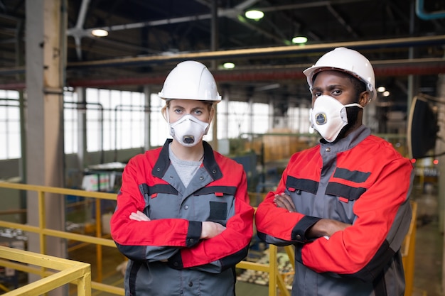 Porträt von ernsthaften Arbeitern in Atemmasken und Schutzhelmen, die mit verschränkten Armen auf Rahmenbrücke im Fabrikladen stehen