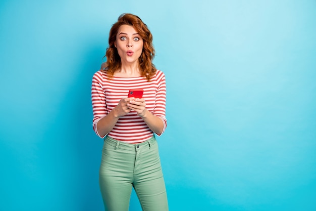 Porträt von erstaunten Mädchen verwenden Handy lesen soziale Netzwerkinformationen beeindruckt schreien schreien wow omg tragen gut aussehen Outfit isoliert über blaue Farbe