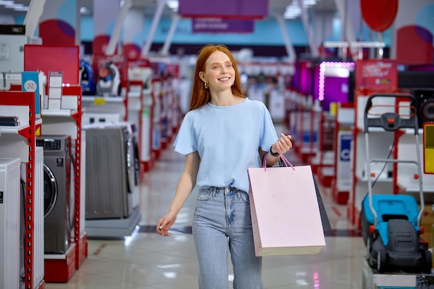 Porträt von Frauen, die im Laden durch Haushaltsgeräte gehen, Taschen halten, lächeln, erfolgreich einkaufen, allein. Rothaarige schöne Dame in Freizeitkleidung am schwarzen Freitag, auf der Suche nach Rabatten