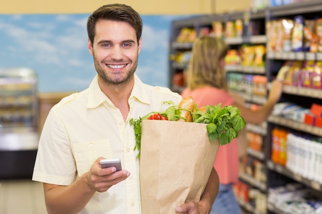 Porträt von lächelnden Lebensmittelprodukten des gutaussehenden Mannes kaufend und seinen Smartphone verwendend