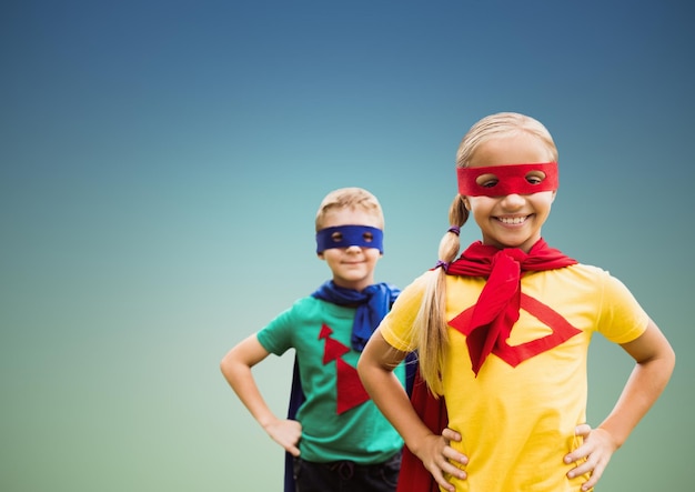 Porträt von lächelnden Superkindern in Umhang und Maske, die mit der Hand auf der Hüfte gegen den klaren Himmel stehen