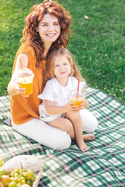 Porträt von Mama und ihrem Kind, das Plastikbecher mit Orangensaft zeigt, die auf der Decke im Park sitzen