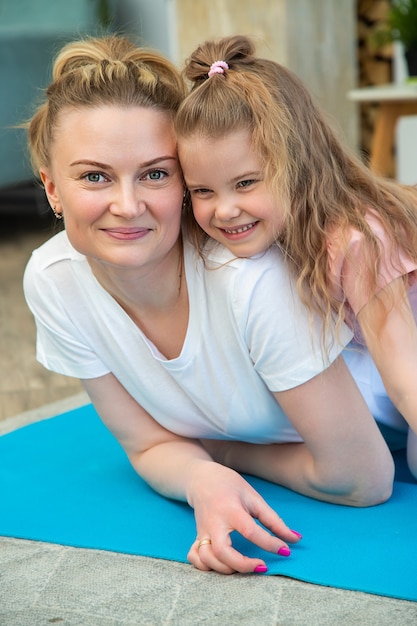Porträt von Mutter und Tochter in Sportkleidung, die auf einer Yogamatte liegen, sich umarmen und in die Kamera schauen.