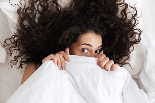 Porträt von oben von der niedlichen Frau 20s mit dunklem lockigem Haar, das ihr Gesicht mit weißer Decke bedeckt, während sie nach dem Aufwachen am Morgen im Bett liegt