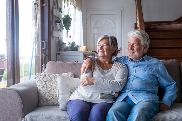 Porträt von Paaren von zwei glücklichen und gesunden Senioren, die das Fenster lächeln und betrachten. Nahaufnahme von reifen Großeltern, die zu Hause auf dem Sofa drinnen Spaß haben und Spaß haben.