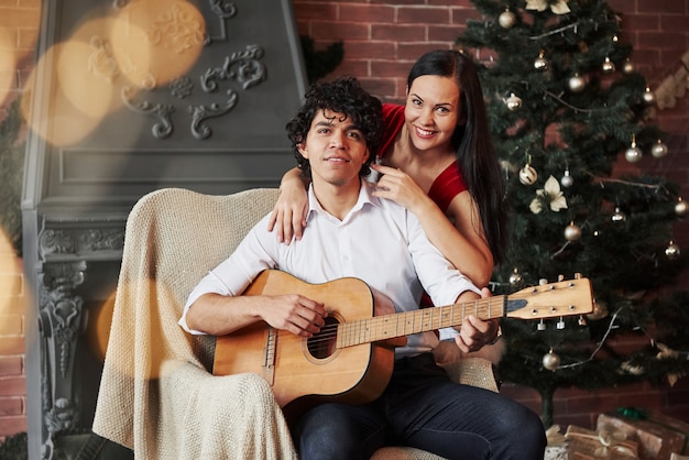 Porträt von reizenden paaren an den feiertagen. gelockter behaarter attraktiver kerl, der auf dem stuhl mit akustikgitarre mit weihnachtsbaum hinten sitzt. freundin im weißen kleid umarmt ihren freund