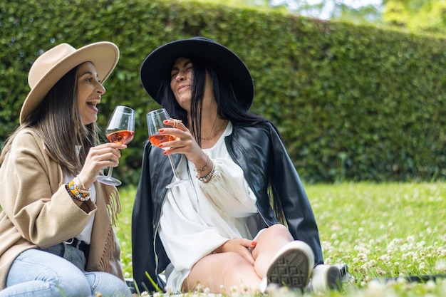 Porträt von schönen lächelnden Frauen mit Hüten, die im Park Wein trinken und anstoßen