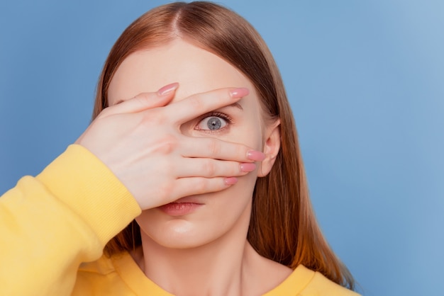 Porträt von verrückten, schockierten, ängstlichen Damenhänden bedecken den Blick, der durch die Finger auf blauem Hintergrund blickt