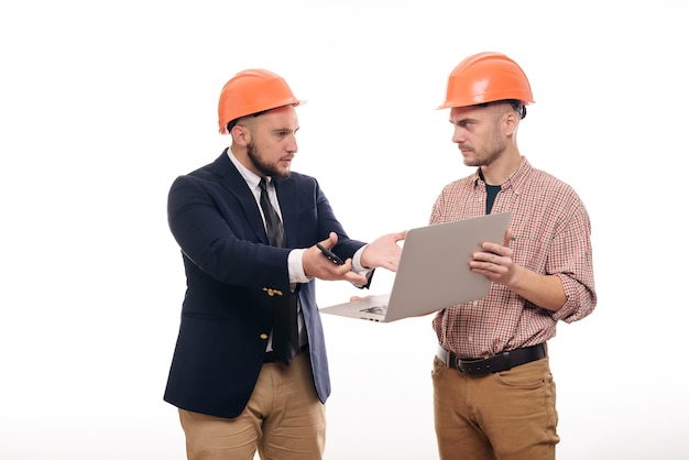 Porträt von zwei Bauherren in den schützenden orange Helmen, die auf weißem lokalisiertem Hintergrund stehen und Laptopanzeige betrachten. Bauprojekt besprechen