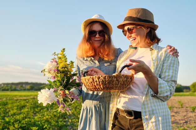 Porträt von zwei glücklich lächelnden umarmten Frauen, Mutter und Tochter im Teenageralter mit Korb mit Bauerneiern in Hüten, mit Blumenstrauß im Garten, Sommernatur, Sonnenuntergangshintergrund