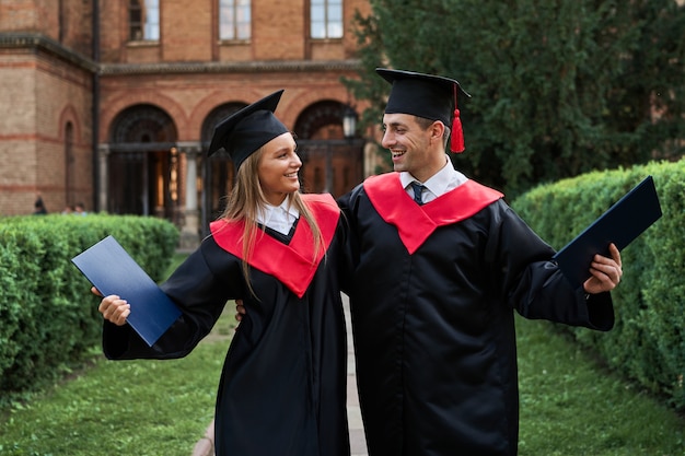 Porträt von zwei graduierten Freunden in Abschlussroben, die auf dem Universitätscampus gehen und sprechen.