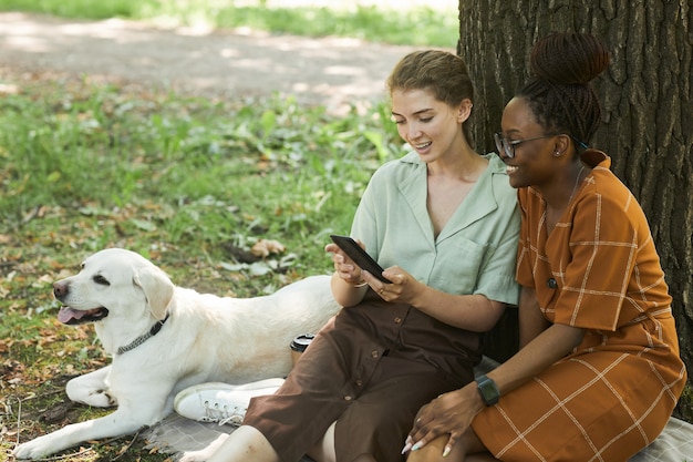 Porträt von zwei jungen Frauen, die auf grünem Gras im Park mit Labrador-Hund sitzen und Smartphone verwenden, Kopierraum