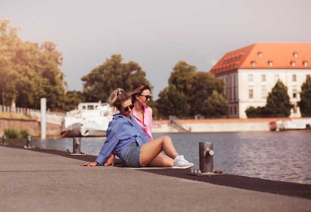 Porträt von zwei jungen, schönen, lächelnden Hipster-Frauen in trendiger Sommerfarbe kleidet sorglose Frauen, die auf Flusshintergrund posieren Positive Models, die Spaß am Umarmen haben