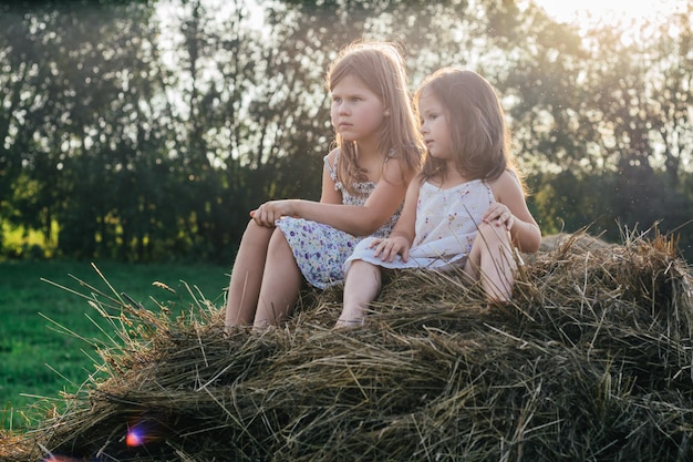 Porträt von zwei Kindermädchen, die auf Strohballen im Heufeld sitzen. Heller sonniger Tag. Fröhlich und genießen Sie das Freundschaftskonzept. Frische Luft auf dem Land. Seitenansicht. Bäume im Hintergrund. Sonnenflecken