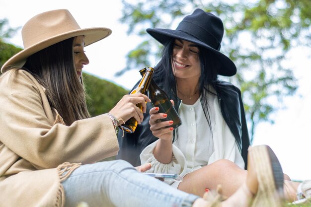 Porträt von zwei lächelnden jungen Frauen mit Hüten, die im Park mit Bierflaschen anstoßen