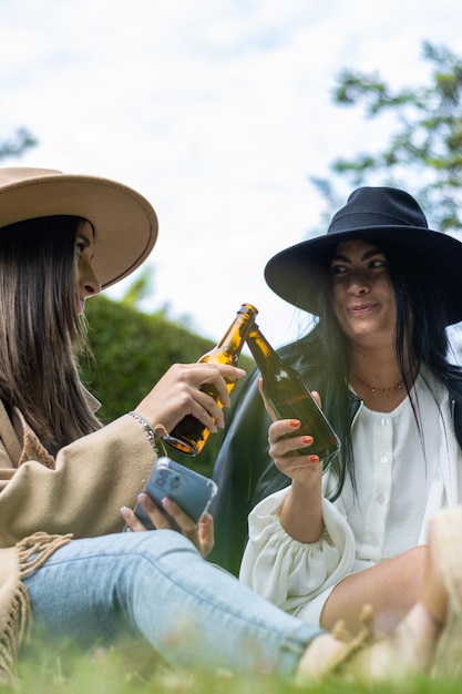 Porträt von zwei lächelnden jungen Frauen mit Hüten, die im Park mit Bierflaschen anstoßen