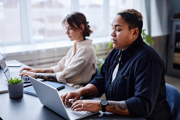 Porträt von zwei modernen Frauen, die Laptops verwenden, während sie gemeinsam im Büro arbeiten