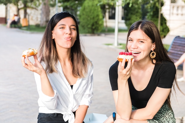 Porträt von zwei schönen Freundinnen, die köstliche Donuts mit Karamell und Himbeere im Stadtpark genießen. Junk aber leckeres Essen für positive Stimmung.