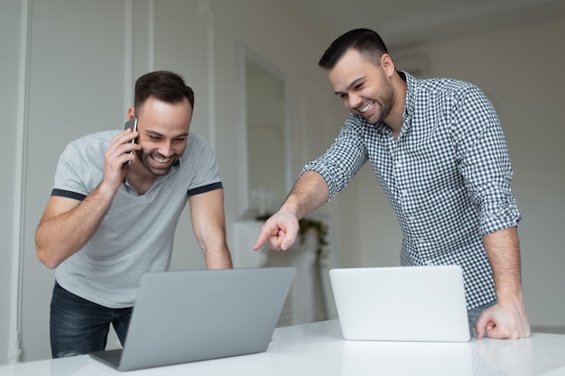 Porträt von zwei selbstbewussten Geschäftsleuten, die am Laptop arbeiten. Ein Mann spricht auf dem Smartphone, wenn ein anderer Mann mit dem Finger auf dem Bildschirm zeigt.