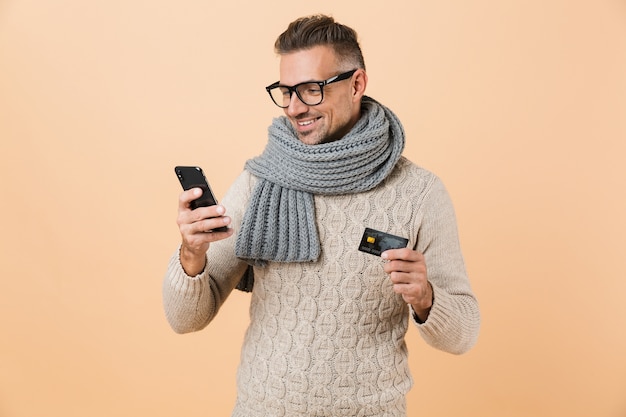 Porträt, wenn ein lächelnder Mann, gekleidet in Pullover und Schal, isoliert über beige Wand steht, Handy hält, Kreditkarte zeigt