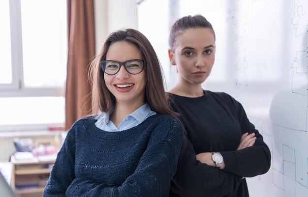 Porträt zweier junger Studentinnen, die vor einer weißen Tafel stehen und in die Kamera schauen