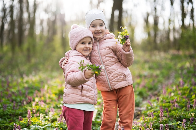 Porträt zweier kleiner Schwestern mit Blumenstrauß im sonnigen Wald Frühlingsfreizeitkonzept im Freien