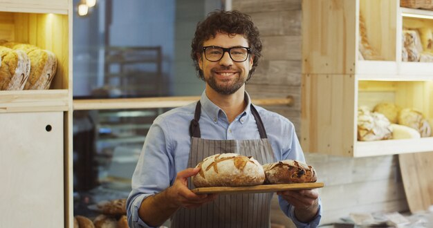 Porträtaufnahme des attraktiven jungen Mannes, Bäckereiverkäufer, der ein Tablett mit frischem Brot hält und mit einem Lächeln in die Kamera schaut. Drinnen