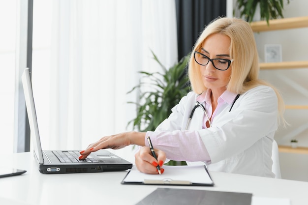Porträtaufnahme einer Ärztin mittleren Alters, die am Schreibtisch sitzt und in der Arztpraxis arbeitet